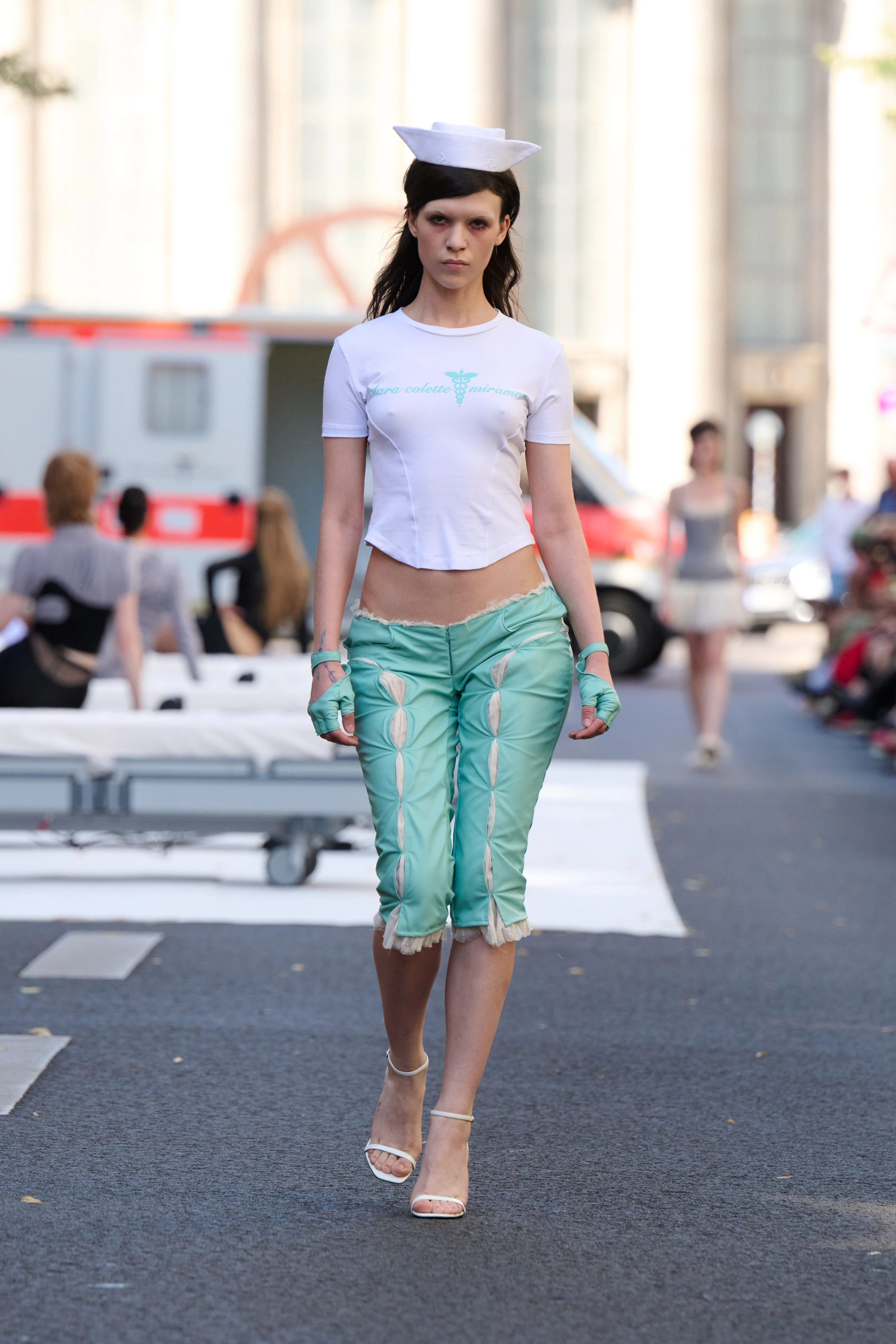 White shirt and turquoise capri pants from Clara Colette Miramon SS26 collection at Berlin Fashion Week worn by a model. Styled with a cap reminiscent of nurses from the 60s.