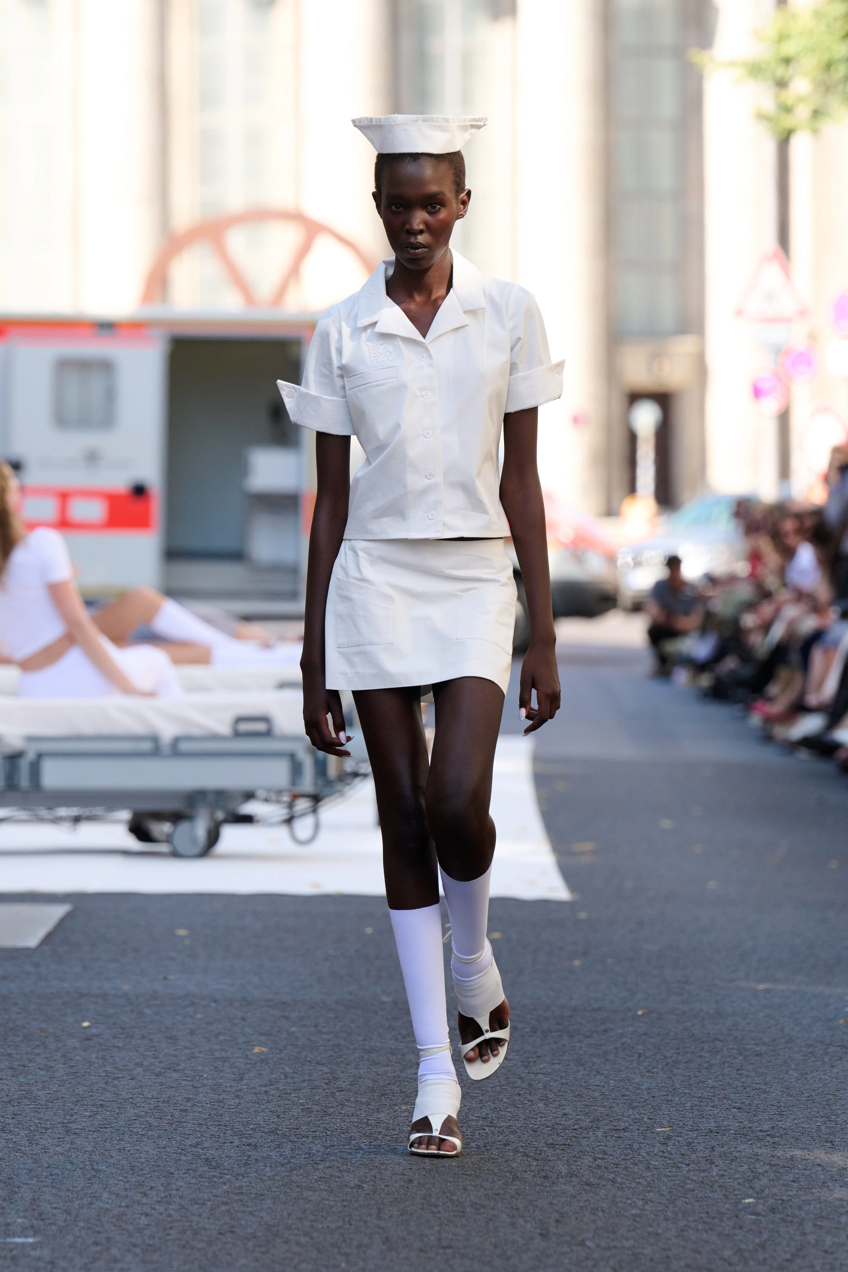 White blouse and skirt from Clara Colette Miramon SS26 collection at Berlin Fashion Week worn by a model. Styled with knee-high socks and a cap reminiscent of nurses from the 60s.