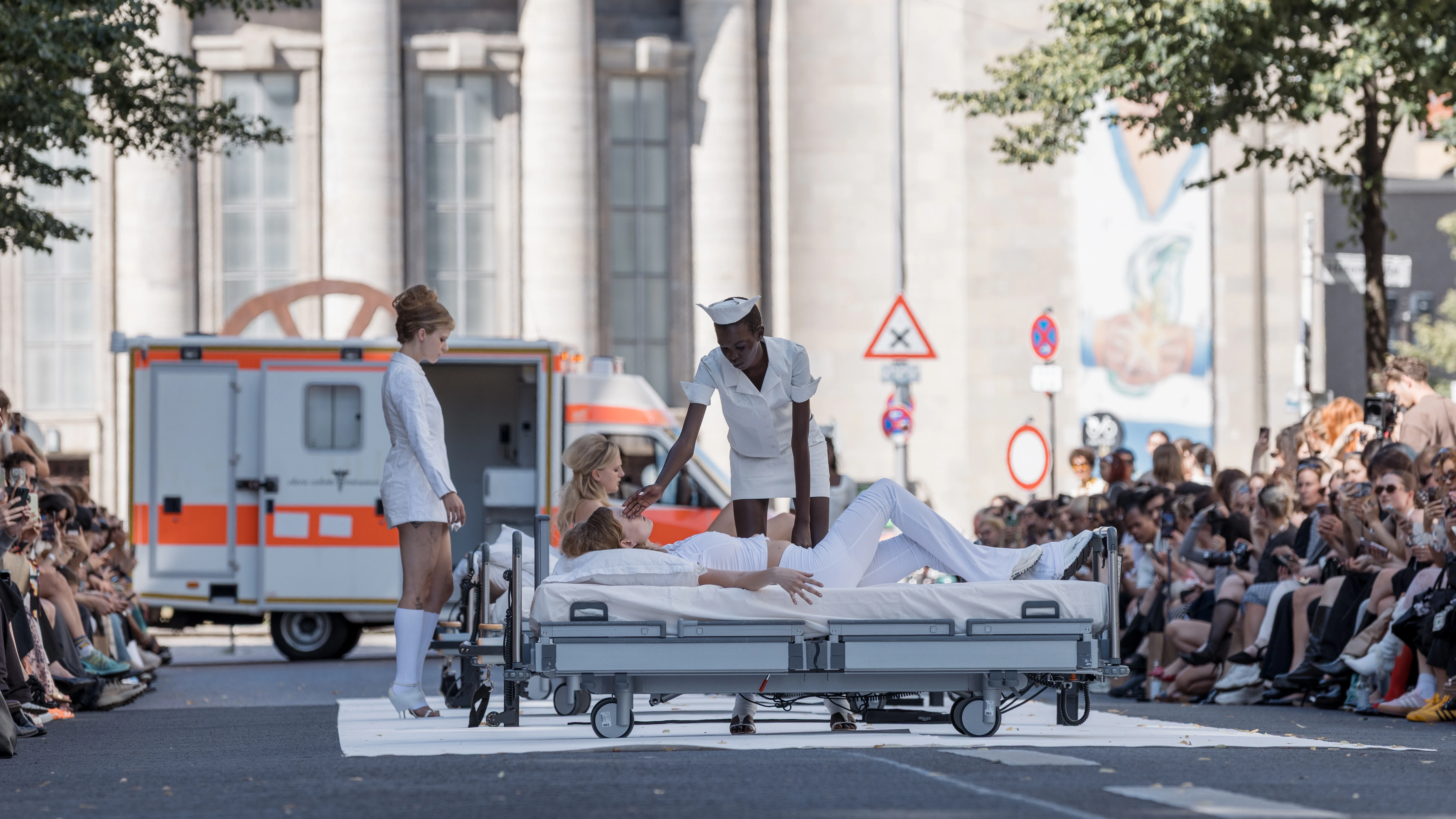 Models at Clara Colette Miramon SS26 show at Berlin Fashion Week whose outfits are reminiscent of hospital nurses caring for another model lying on a hospital bed.